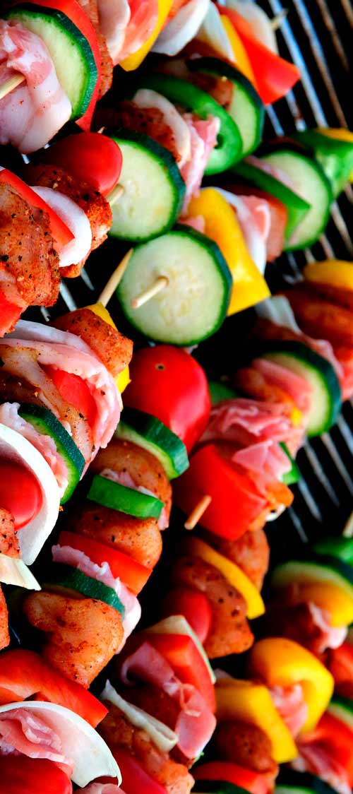 Spiedini colorati su una griglia, con carne, peperoni rossi e gialli, cipolla bianca, zucchine e pomodorini.