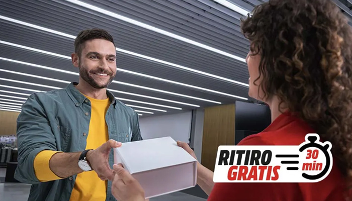 Un uomo consegna una scatola bianca a una donna. Un cartello indica "Ritiro Gratis 30 min". Lo sfondo è un soffitto con luci lineari.