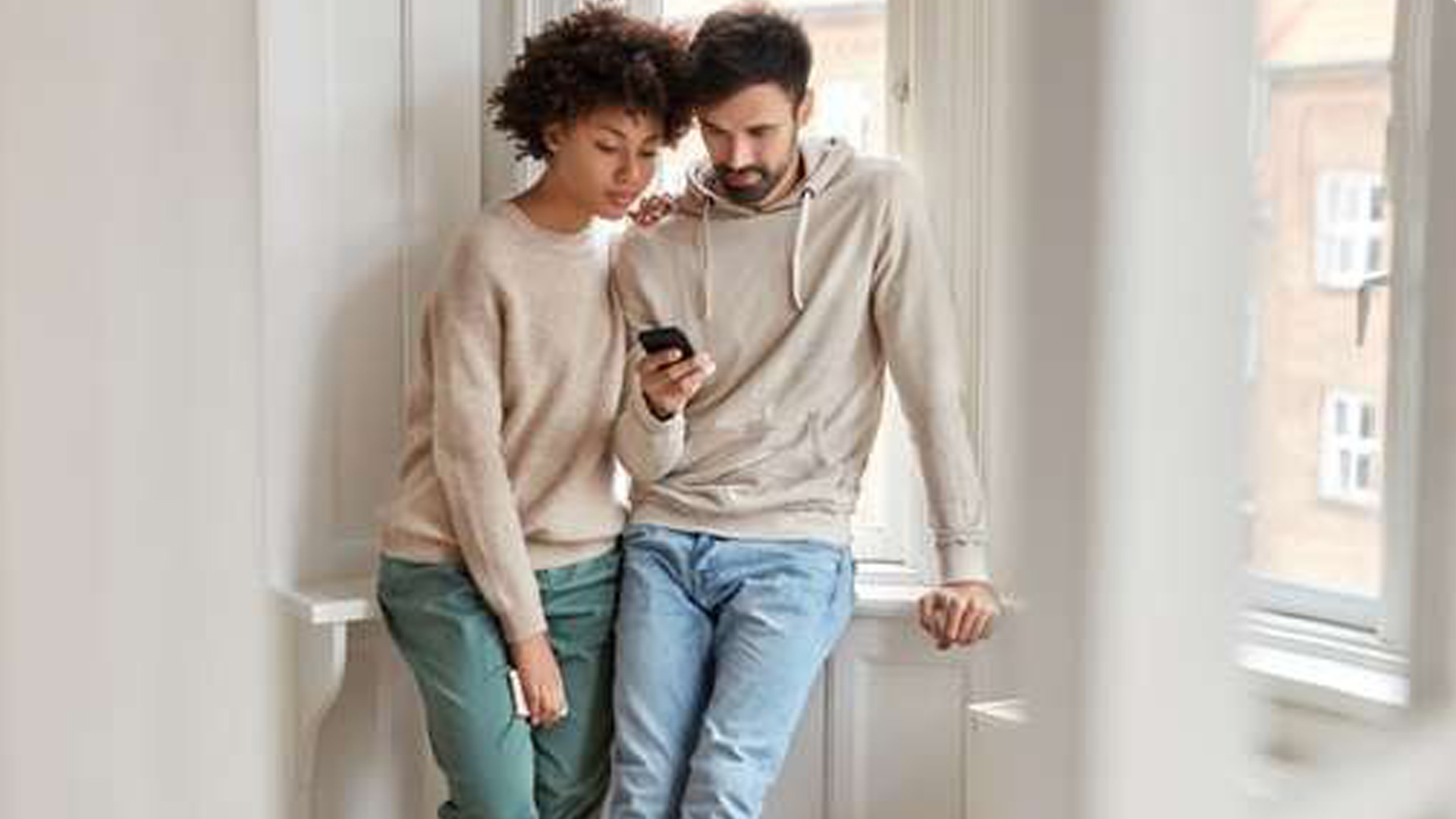 Uomo e donna guardano un telefono vicino alla finestra. L'uomo indossa una felpa beige. La donna indossa un maglione beige.
