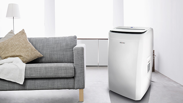 Climatizzatore mobile KOENIC KAC 14022 WLAN in bianco accanto a un divano grigio in un soggiorno moderno. 