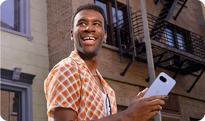 Un uomo sorridente tiene un telefono cellulare. Sullo sfondo, un edificio in mattoni con balconi in metallo.