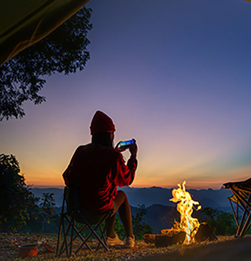 Persona seduta su una sedia pieghevole che fotografa un fuoco da campo al tramonto con montagne sullo sfondo.