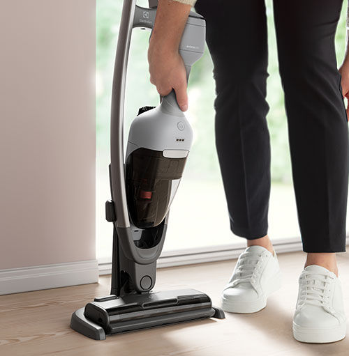 Persona usa aspirapolvere verticale grigio su pavimento di legno, vicino a finestra. Indossa pantaloni neri e scarpe da ginnastica bianche.