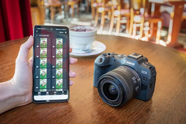 Su un tavolo di legno sono presenti un telefono che mostra foto, una fotocamera Canon e una tazza di caff�.