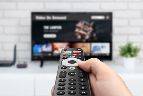 Mano che tiene un telecomando nero puntato verso una TV che mostra un'interfaccia di streaming su un mobile bianco.