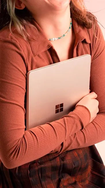 Donna che tiene un portatile color crema con un logo Windows nero. Indossa una maglia color zucca, una gonna a quadri e una collana.