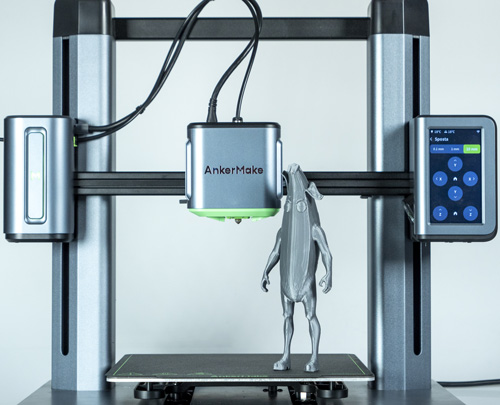 Stampante 3D in argento con schermo a destra, stampa una figura grigia su una piattaforma nera.