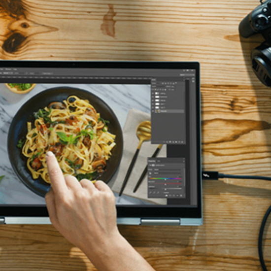 Schermo di laptop su tavolo di legno con foto di pasta in un piatto nero. Una mano tocca lo schermo; un controller � in alto a destra.