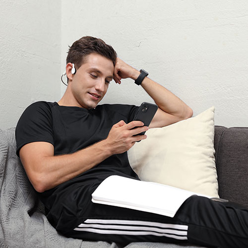 Uomo seduto su un divano, indossa cuffie e guarda un telefono. C'� un cuscino e un libro bianco.