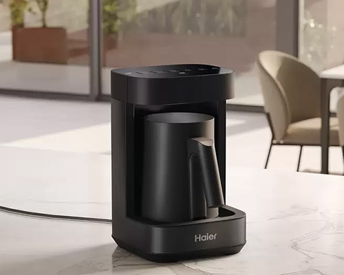 Una macchina da caff� nera marca Haier su un piano bianco, con una sedia e una finestra sullo sfondo.