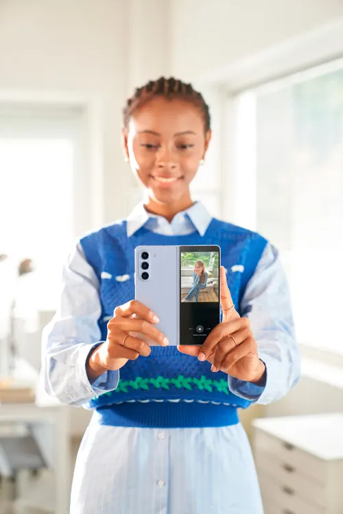 Donna con maglia blu tiene in mano un telefono pieghevole aperto, con tre fotocamere sul retro. Sullo schermo si vede una persona.