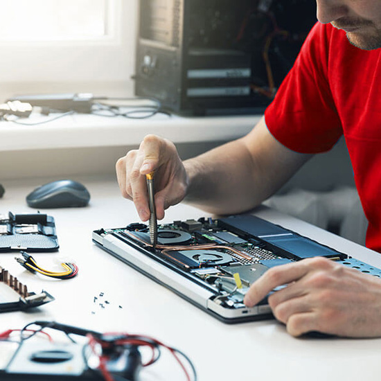 Uomo con maglietta rossa ripara un laptop aperto con cacciavite. Sul tavolo, componenti elettronici e un computer fisso sullo sfondo.
