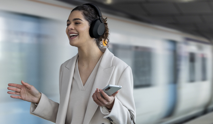 Donna sorridente con cuffie nere, giacca chiara, telefono in mano. Treno in movimento sullo sfondo.