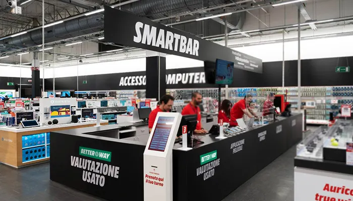 Interno di un negozio di elettronica con bancone "Smartbar" nero, insegne bianche, e personale in magliette rosse. Espositori con dispositivi.