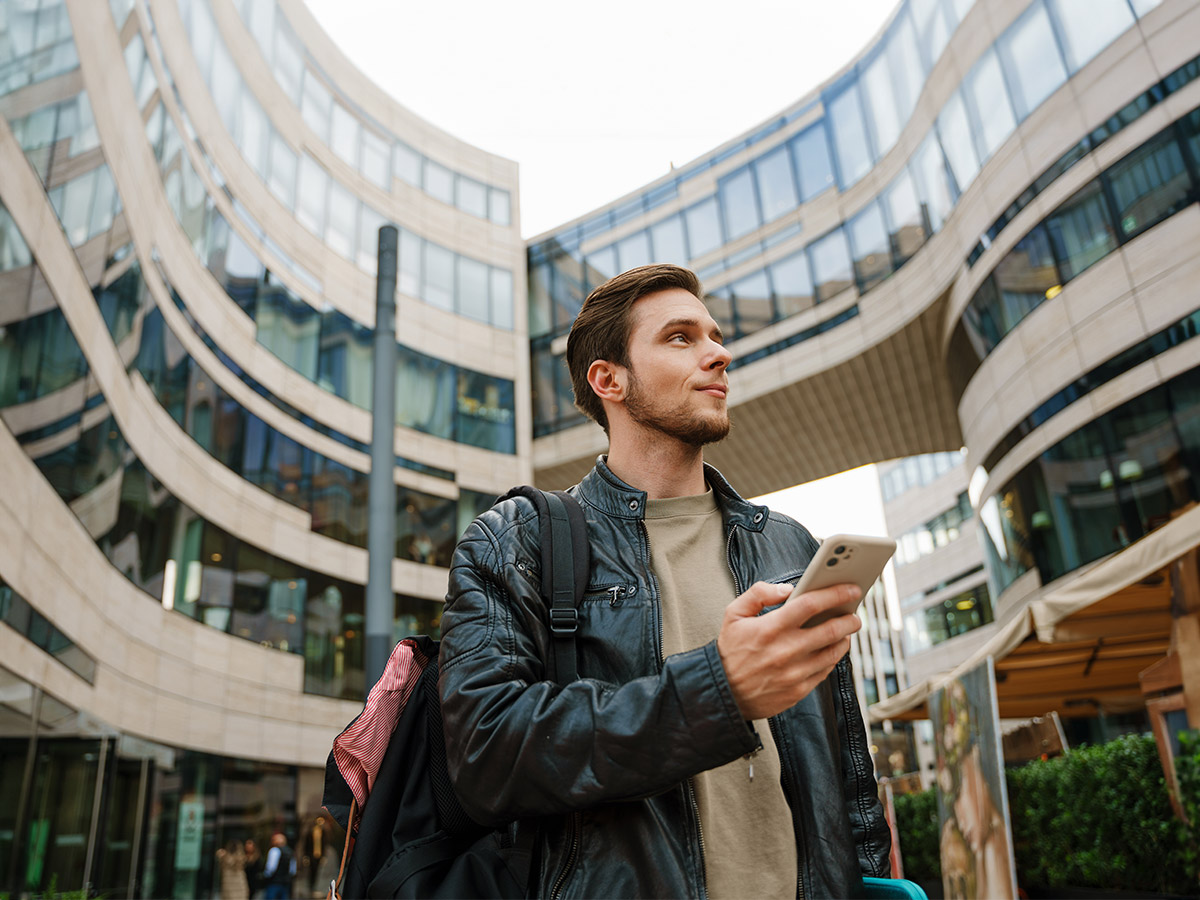 Uomo con giacca di pelle nera e zaino, tiene un telefono. Sullo sfondo, un edificio moderno con vetrate.