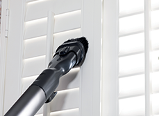 Aspirapolvere nero pulisce persiane bianche. Le lamelle sono orizzontali e uniformemente distanziate. Sfondo luminoso.