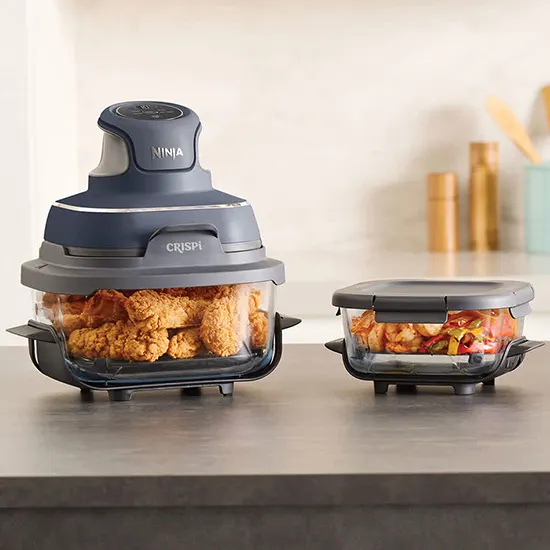 Friggitrice ad aria Ninja Crispi in vetro blu scuro con pollo fritto all'interno, posizionata accanto a un secondo contenitore ermetico con gamberi e peperoni sul bancone.