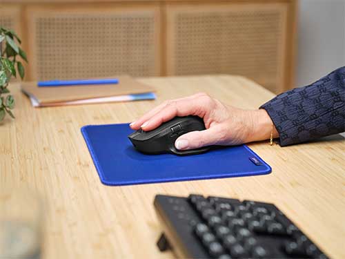 Una mano utilizza un mouse nero su un tappetino blu. Una tastiera è in primo piano su una scrivania di legno, con un notebook sullo sfondo.