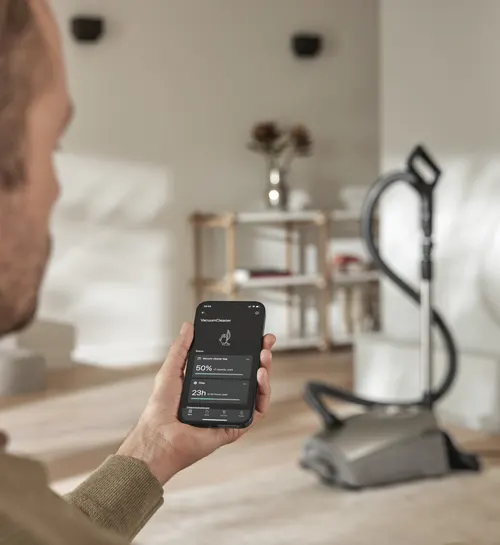 Uomo con telefono che mostra app di aspirapolvere robot. Aspirapolvere robot sullo sfondo in una stanza con scaffali.