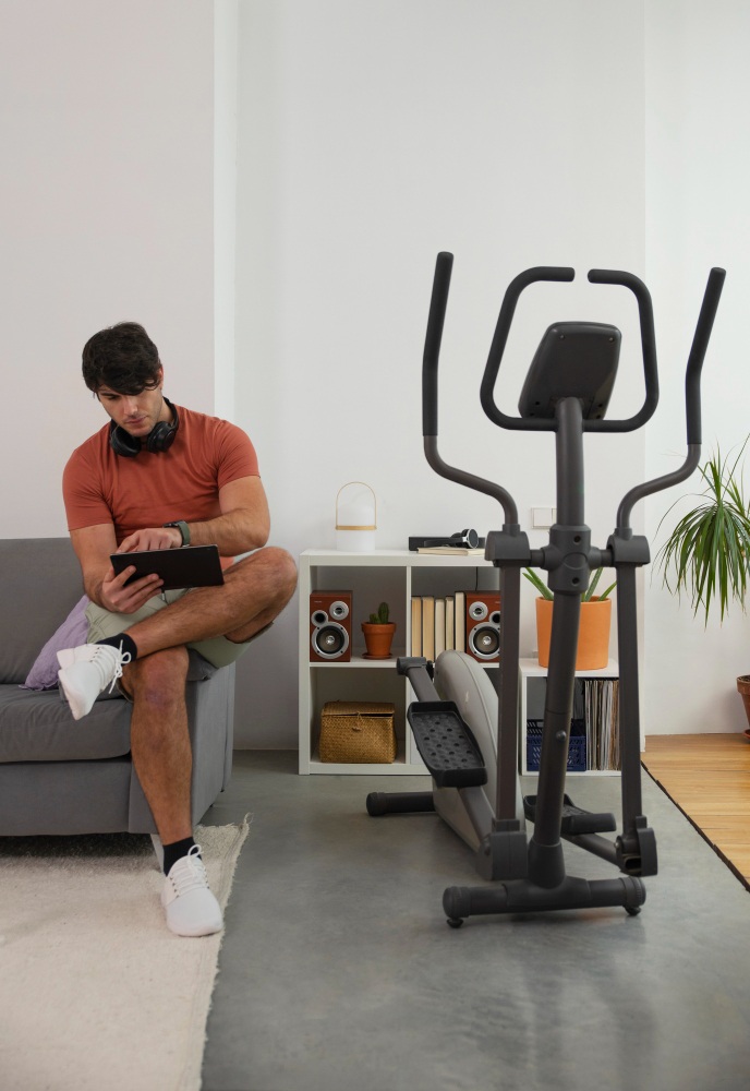 Uomo seduto su divano grigio con tablet e cuffie. Dietro, mobile bianco con oggetti e ellittica nera su tappeto grigio. Pianta.
