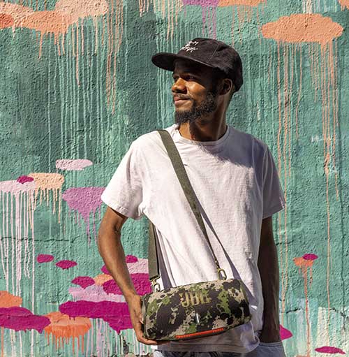 Uomo con cappello e maglietta bianca, porta una borsa JBL a tracolla davanti a un muro colorato.