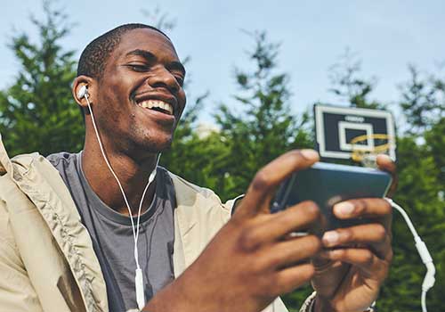 Uomo sorridente con cuffie, guarda uno smartphone. Dietro, alberi e canestro da basket.