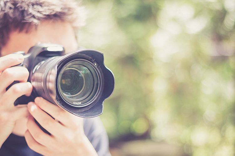 Un uomo tiene una fotocamera nera con un grande obiettivo. Lo sfondo � sfocato con colori verdi e marroni.
