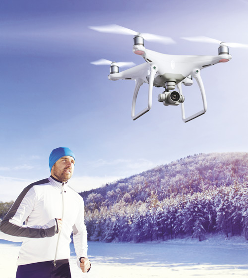 Uomo che corre in un campo innevato con un drone bianco che vola sopra. Alberi e cielo azzurro sullo sfondo.