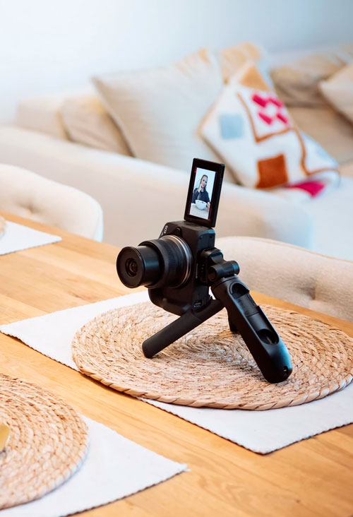 Fotocamera nera su treppiede, su un tavolo di legno con tovagliette. Divano chiaro con cuscini sullo sfondo.