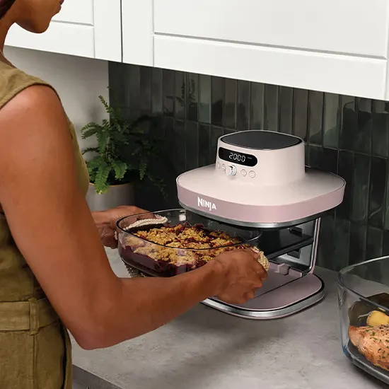 Una donna inserisce una pirofila in vetro con un crumble di frutta nella friggitrice ad aria Ninja di colore rosa chiaro.