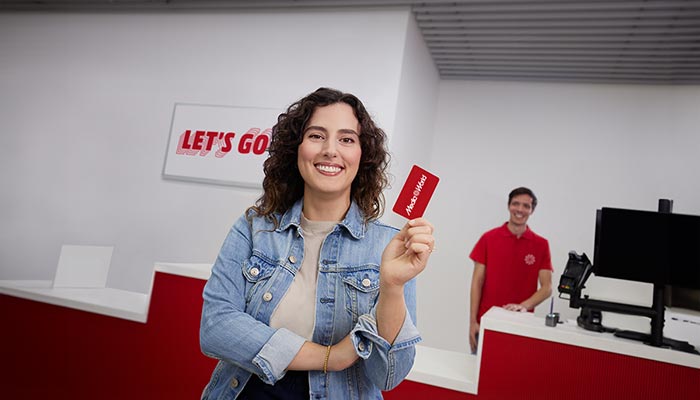 Donna sorridente mostra tessera rossa. Insegna "Let's Go" e bancone rosso-bianco sullo sfondo. Uomo in maglietta rossa dietro al bancone.