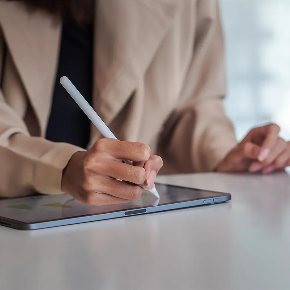 Una persona usa una penna digitale su un tablet grigio, con una giacca beige su un tavolo bianco.