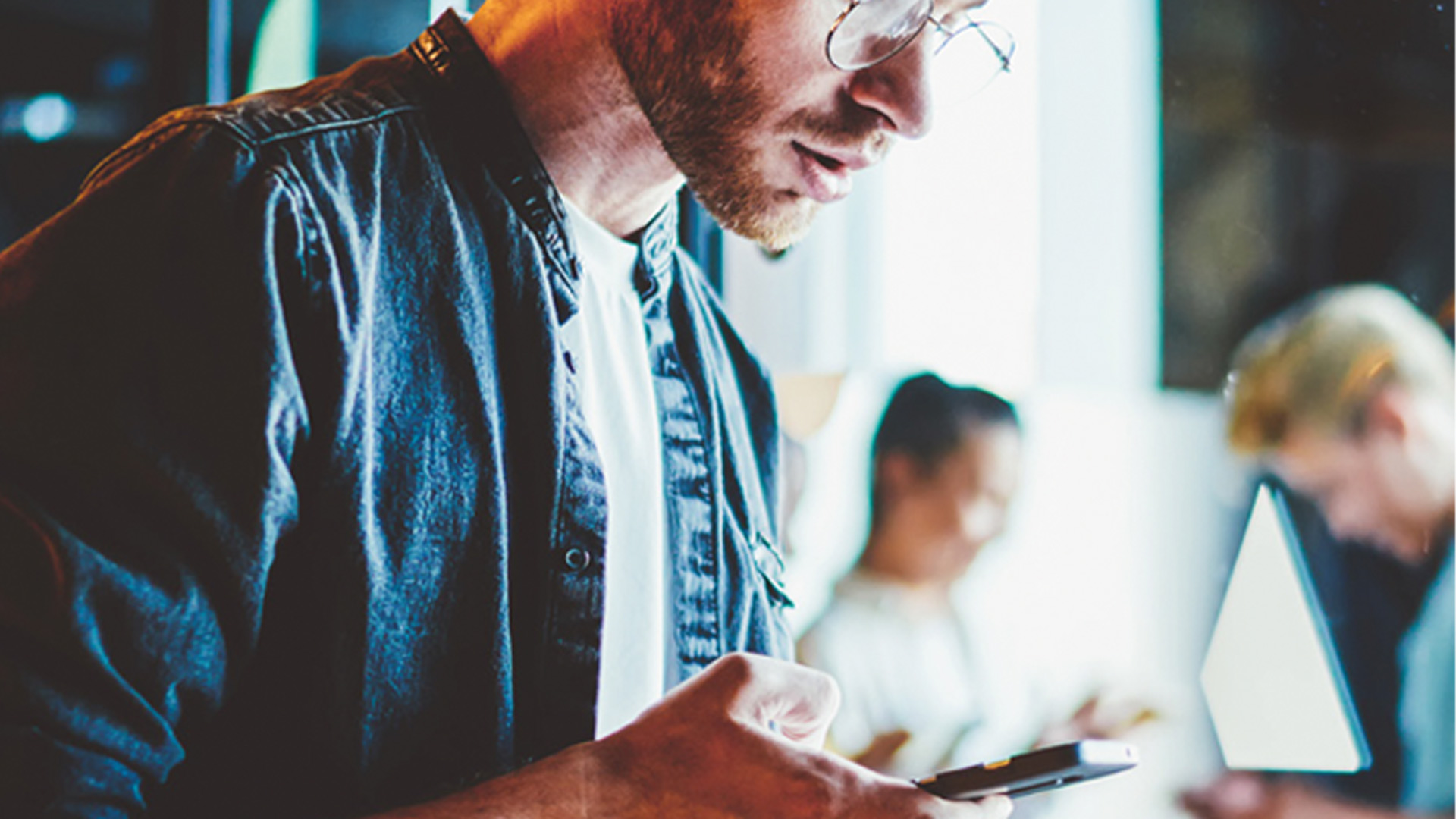 Un uomo con gli occhiali usa un telefono. Indossa una camicia blu su una t-shirt bianca. Altre persone sono sullo sfondo.