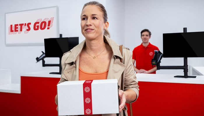 Donna tiene una scatola bianca con nastro rosso in negozio. Un uomo in rosso è sullo sfondo, vicino a monitor neri.