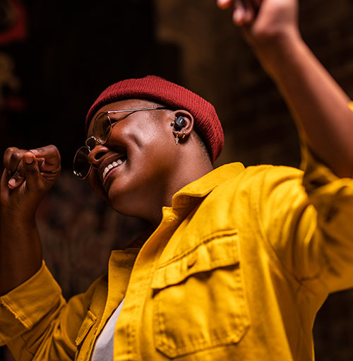 Persona sorridente con berretto rosso e camicia gialla. Indossa occhiali e auricolare. Braccia alzate su sfondo scuro.