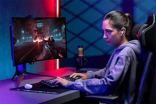 Ragazza con cuffie gioca con computer su sedia gaming. Schermo mostra gioco d'azione, microfono dorato sul tavolo. Luci blu e rosse.