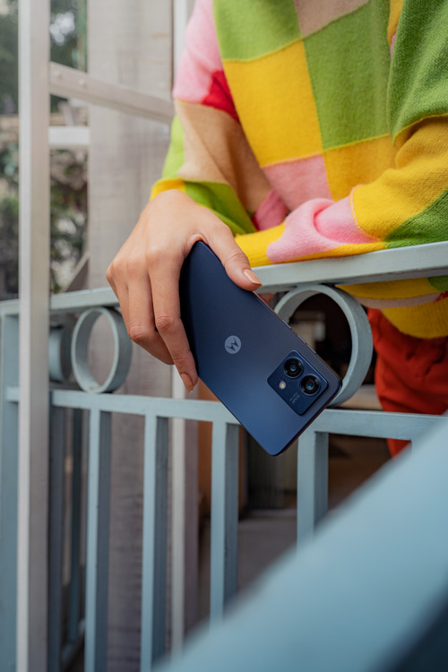 Una mano tiene un telefono blu contro una ringhiera bianca. La persona indossa una maglia a scacchi colorata.