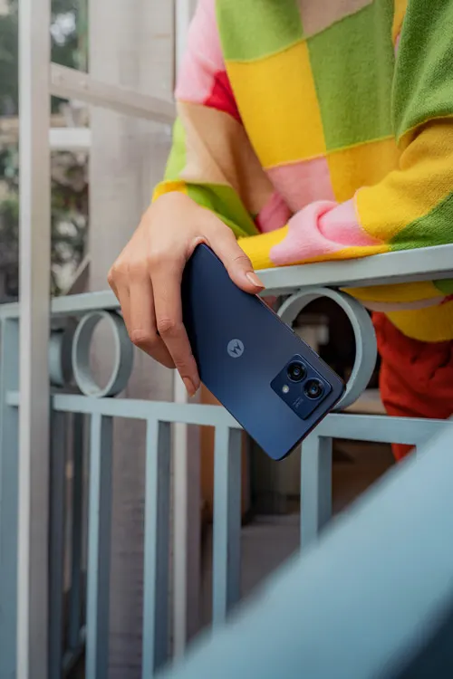 Una mano tiene un telefono blu contro una ringhiera bianca. La persona indossa una maglia a scacchi colorata.