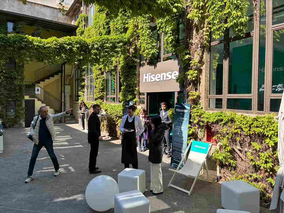 Evento Hisense all'aperto in un cortile con edera, persone in piedi, pouf bianchi e cartelli pubblicitari.