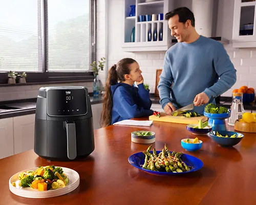 Cucina luminosa con uomo che taglia verdure, ragazza seduta e friggitrice ad aria nera sul tavolo con cibo pronto.