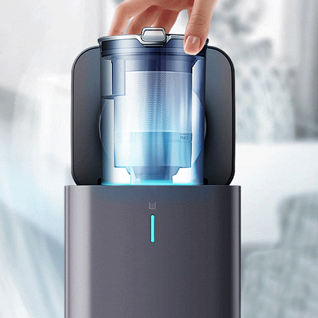 Una mano solleva un serbatoio trasparente da un dispositivo grigio. Il serbatoio ha un coperchio argentato. Una luce blu illumina il dispositivo.