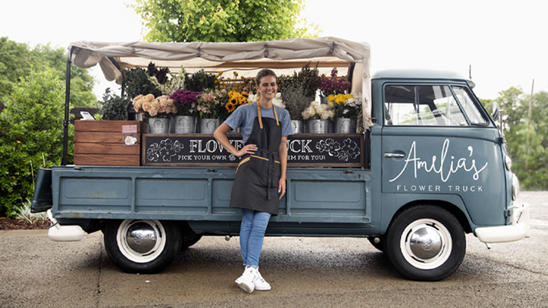 Una donna in grembiule posa davanti a un furgone di fiori blu con fiori in vasi sul retro.