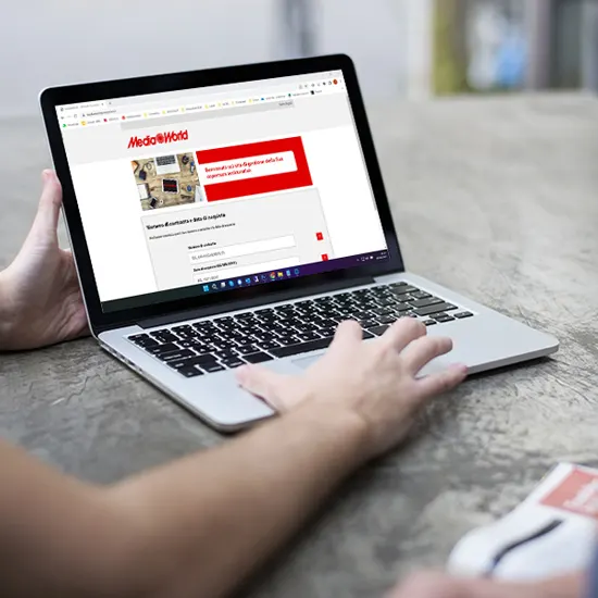 Un laptop aperto mostra il sito MediaWorld. Una mano digita sulla tastiera. Sul tavolo c'è una brochure.