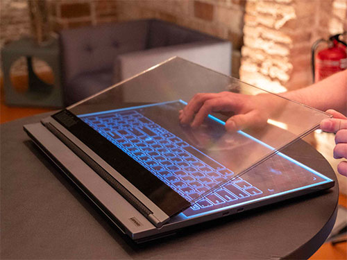 Un computer portatile aperto su un tavolo mostra una tastiera blu illuminata e un pannello di vetro trasparente.