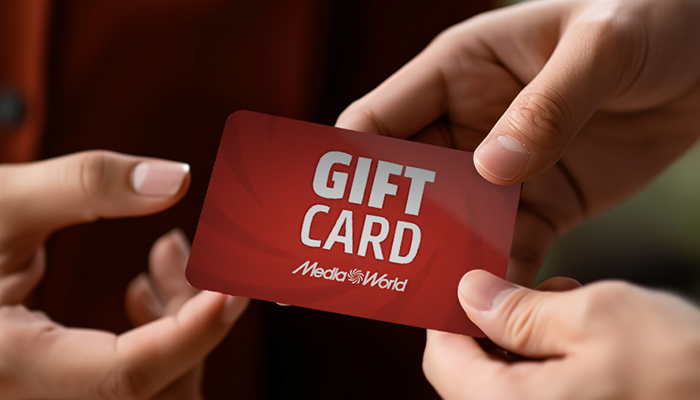 Mani che scambiano una carta regalo rossa con testo "Gift Card" e logo "Media World" su sfondo sfocato.