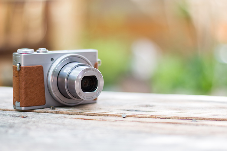Fotocamera digitale argento e marrone su una superficie di legno chiara, con sfondo sfocato di verde e marrone.