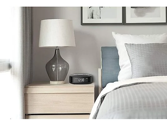 Lampada grigia su un comodino beige con una sveglia digitale. Sullo sfondo, un letto con cuscini.