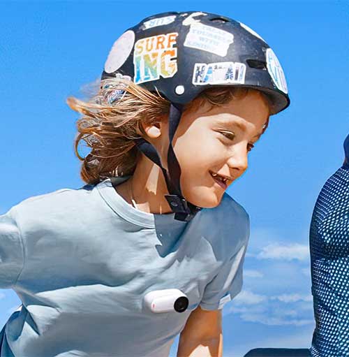 Ragazzo sorridente con casco decorato e maglietta blu, con action camera bianca, su sfondo cielo azzurro.