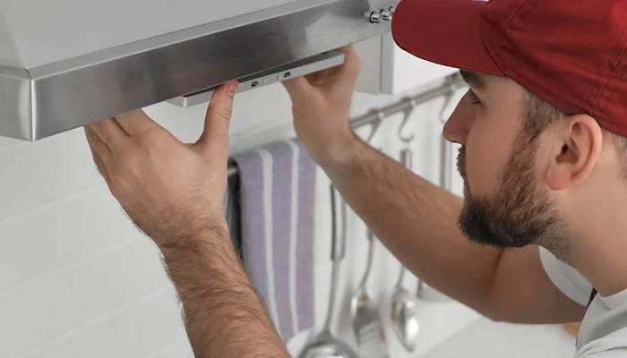 Uomo con cappello rosso che ispeziona una cappa in acciaio inossidabile. Sullo sfondo utensili da cucina appesi.