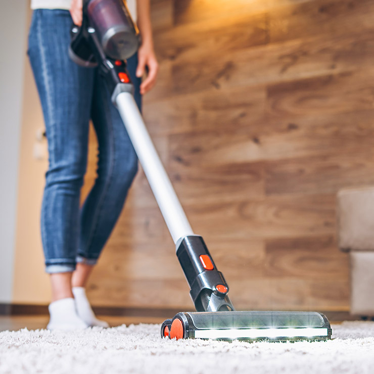 Donna che aspira un tappeto bianco con un aspirapolvere a bastone. Sullo sfondo, pavimento in legno chiaro.